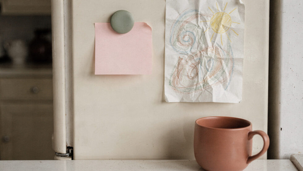 Affirmations for Exhaustion That Come With Something to Do A blank pink note held by a sage magnet on a cream fridge beside a crayon drawing, with a terracotta mug below