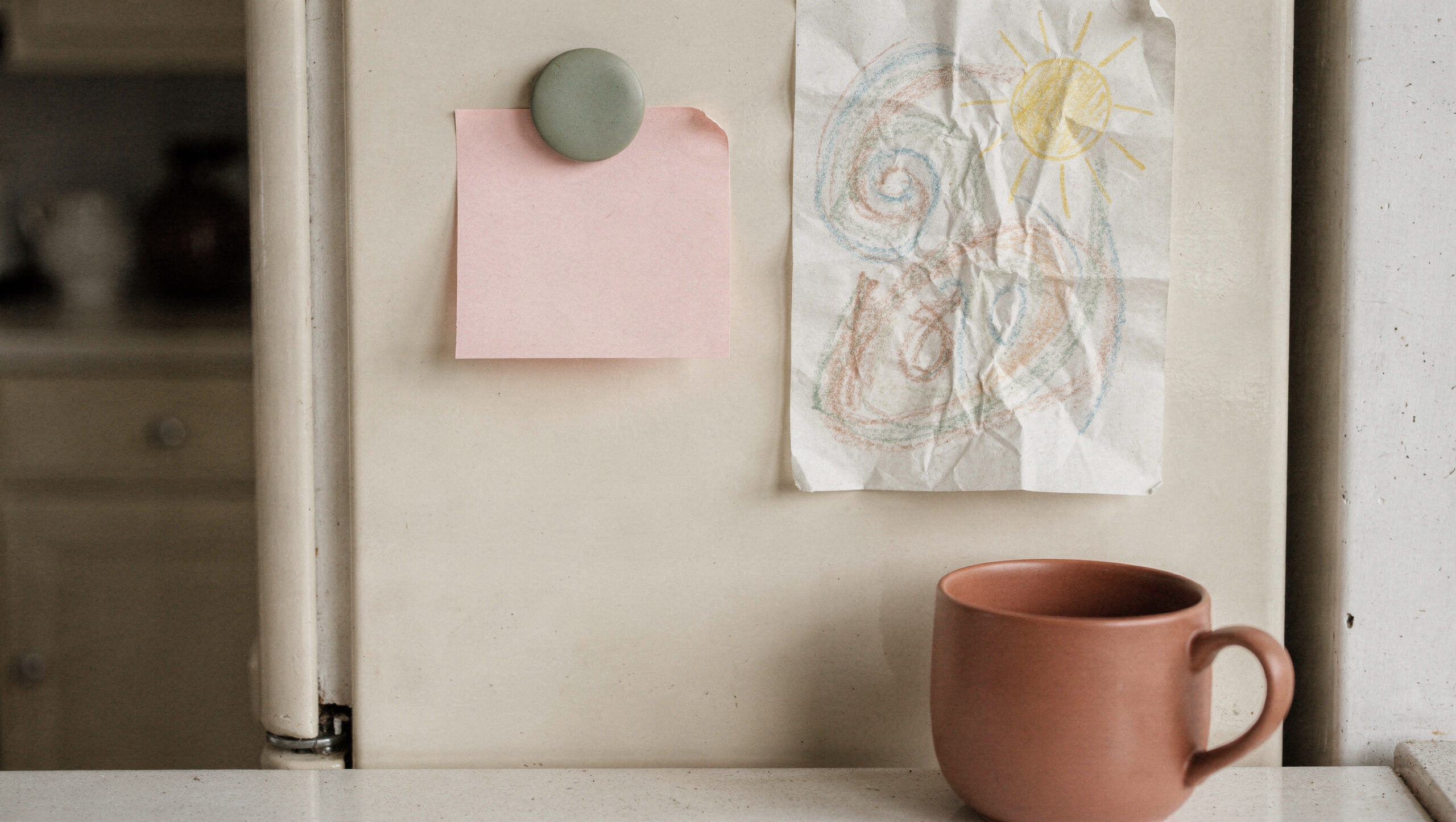 A blank pink note held by a sage magnet on a cream fridge beside a crayon drawing, with a terracotta mug below
