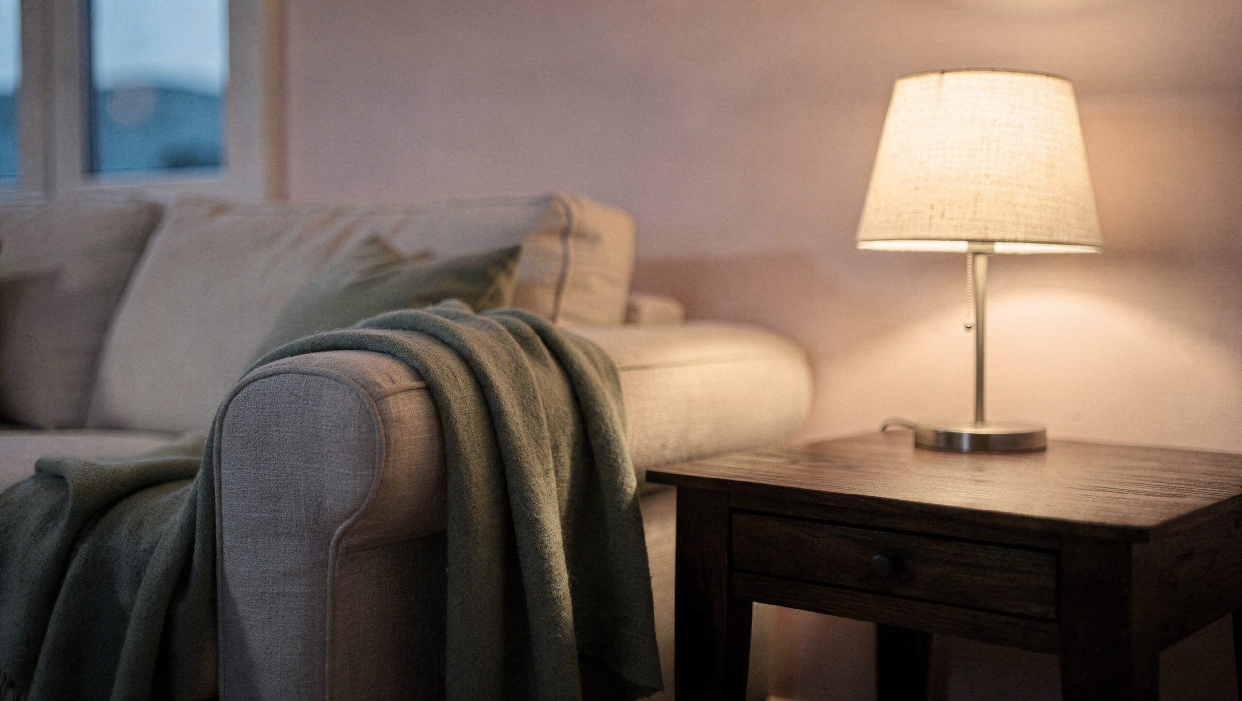A glowing table lamp on a dark wooden side table beside a sofa with a sage throw at dusk