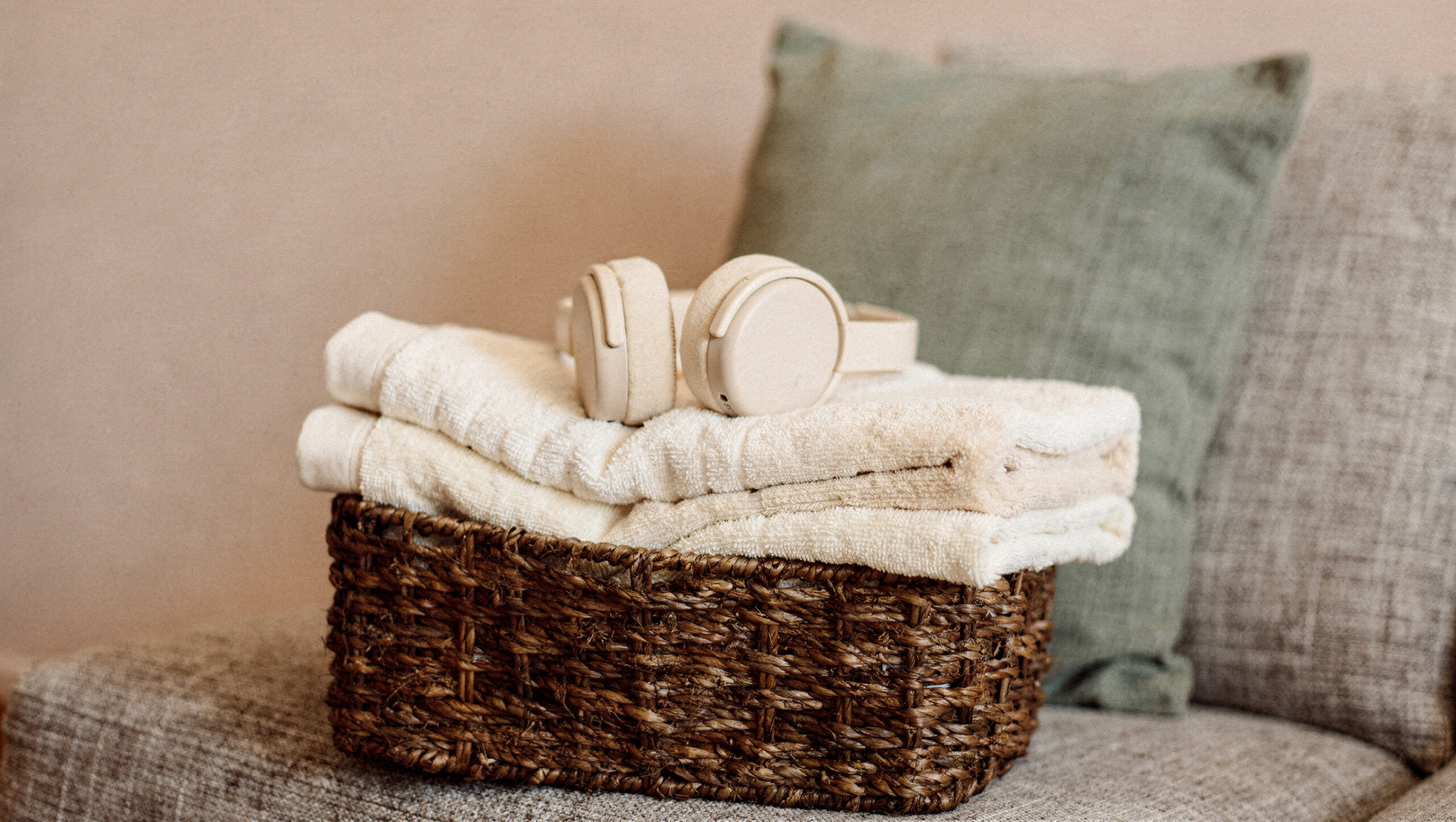 Cream headphones resting on folded cream towels in a woven basket on a sofa with a sage cushion