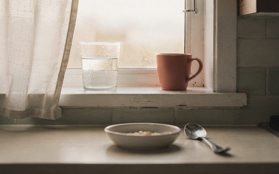A Gentle Morning Routine for Moms Who Can't Wake Up Early A glass of water and a terracotta mug on a kitchen windowsill in pale morning light, a bowl and spoon on the counter