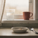 morning-routine-for-moms-who-cant-wake-up-early-featured A glass of water and a terracotta mug on a kitchen windowsill in pale morning light, a bowl and spoon on the counter
