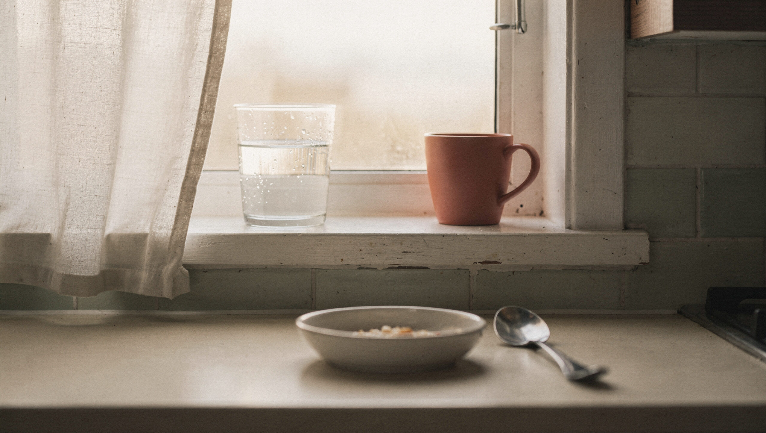 morning-routine-for-moms-who-cant-wake-up-early-featured A glass of water and a terracotta mug on a kitchen windowsill in pale morning light, a bowl and spoon on the counter