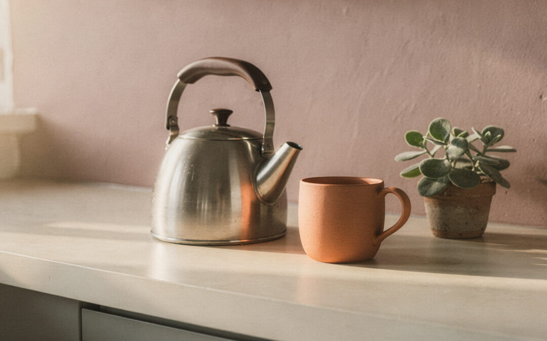 A Small Daily Self-Care Ritual You Can Start Again Tomorrow A kettle, a terracotta mug and a small green plant on a cream counter in morning light