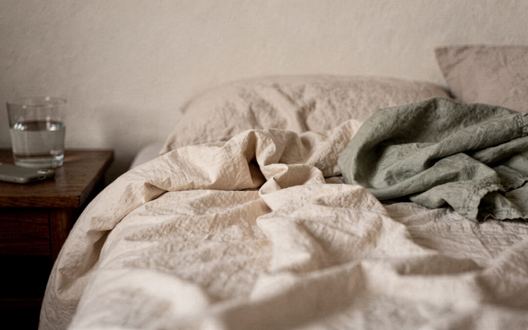 When You Wake Up Already Tired: How to Function on No Sleep as a Mom An unmade bed with a sage blanket beside a nightstand with a glass of water and a phone