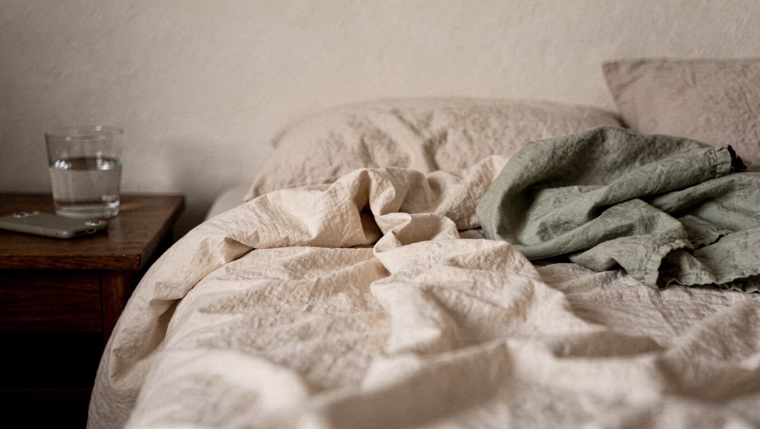 when-you-wake-up-already-tired-featured An unmade bed with a sage blanket beside a nightstand with a glass of water and a phone